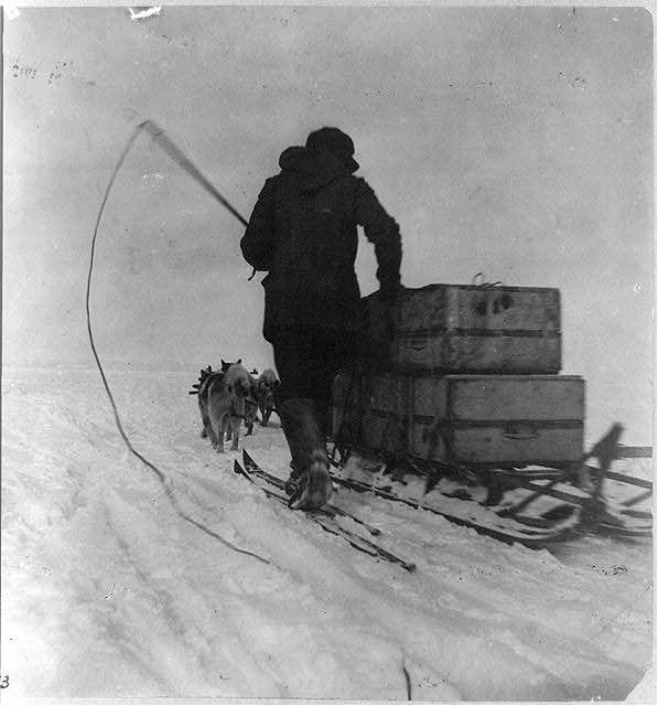 Archival photograph: a man in furs with a whip beside a dog sledge loaded with crates
