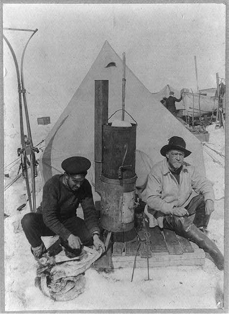 Archival photograph: two men seated at a stove inside a tent